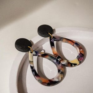 May include: A pair of teardrop earrings with a black circular stud and a colorful, marbled design. The earrings feature a mix of orange, purple, yellow, and white hues, suspended from gold-colored hooks.