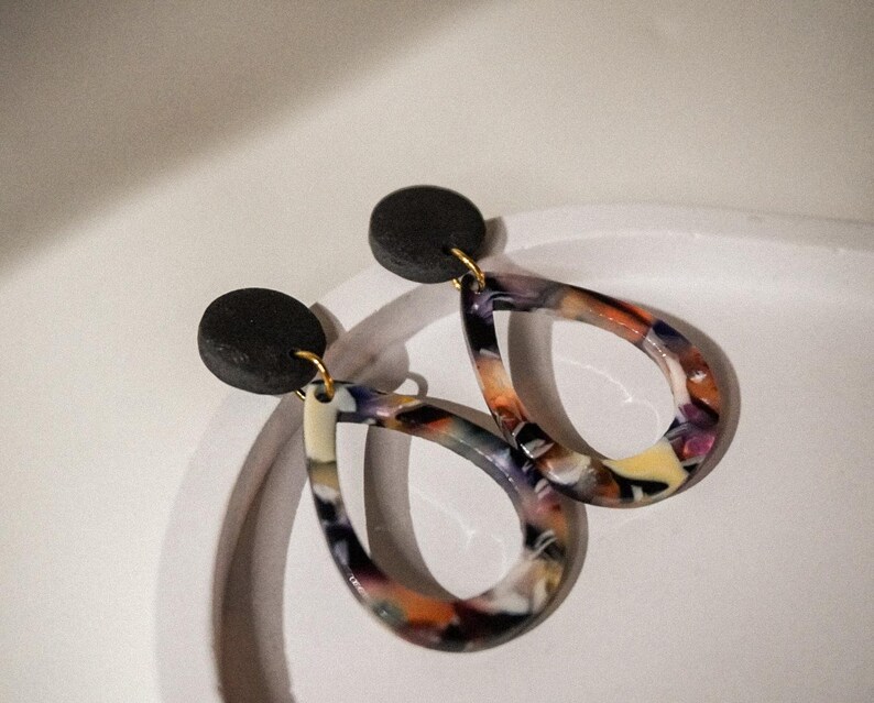 May include: A pair of teardrop earrings with a black circular stud and a colorful, marbled design. The earrings feature a mix of orange, purple, yellow, and white hues, suspended from gold-colored hooks.