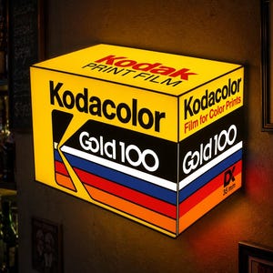 May include: A rectangular illuminated wall sign featuring the Kodak Gold 100 film box design. The yellow sign displays the Kodak logo, the text Kodacolor Film for Color Prints, and Gold 100 with a multi-colored stripe pattern and 35 mm DX text.