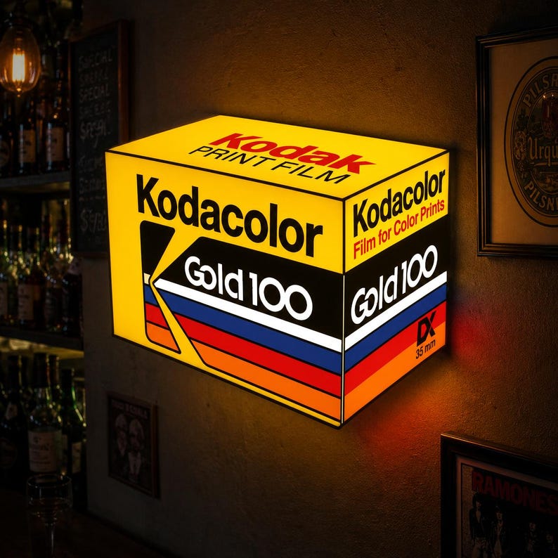 May include: A rectangular illuminated wall sign featuring the Kodak Gold 100 film box design. The yellow sign displays the Kodak logo, the text Kodacolor Film for Color Prints, and Gold 100 with a multi-colored stripe pattern and 35 mm DX text.