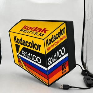 May include: A rectangular lamp shaped like a box of Kodak Gold 100 35mm film. The yellow, black, red, and blue design features the text Kodak Print Film, Kodacolor, Gold 100, and DX 35mm. It includes a black power cord with a small inline switch.