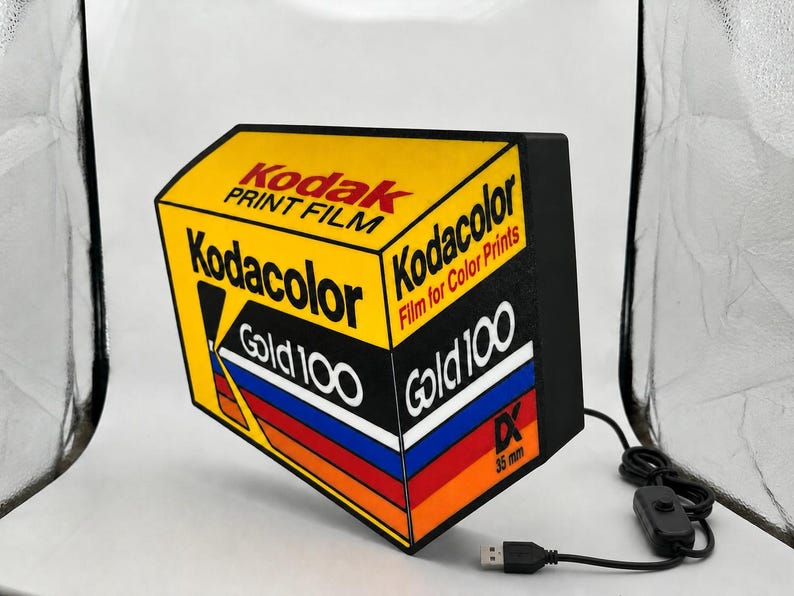 May include: A rectangular lamp shaped like a box of Kodak Gold 100 35mm film. The yellow, black, red, and blue design features the text Kodak Print Film, Kodacolor, Gold 100, and DX 35mm. It includes a black power cord with a small inline switch.