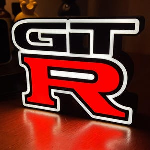 Op de afbeelding: Een zwart, rood en wit GT-R logo bord. De letters GT zijn zwart met witte contouren en de R is rood met een witte contour. Het bord is verlicht en werpt een rode gloed op het oppervlak eronder.