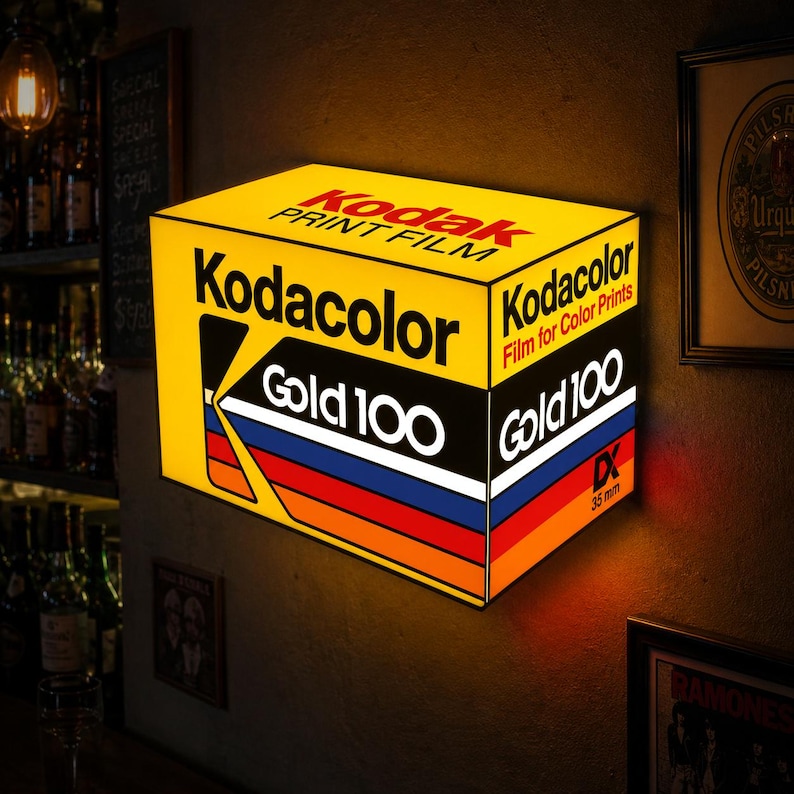 May include: A rectangular illuminated wall sign featuring the Kodak Gold 100 film box design. The yellow sign displays the Kodak logo, the text Kodacolor Film for Color Prints, and Gold 100 with a multi-colored stripe pattern and 35 mm DX text.