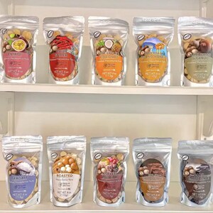 Puede incluir: Exhibición de diez bolsas de plástico transparente llenas de nueces de macadamia con diferentes sabores. Las bolsas están dispuestas en una estantería blanca. Cada bolsa tiene una etiqueta con el nombre del sabor y un logotipo de grano de café. Sabores: maracuyá, café con chile, coco y sal marina.