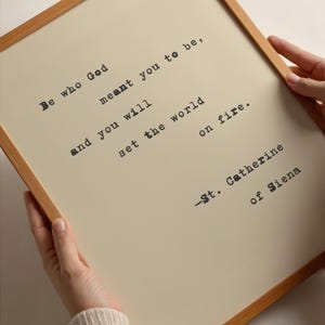 Puede incluir: Una impresión enmarcada con un fondo beige claro y un marco de madera. La impresión presenta una cita de Santa Catalina de Siena en una fuente de máquina de escribir: "Sé quien Dios quiso que fueras, y prenderás fuego al mundo."