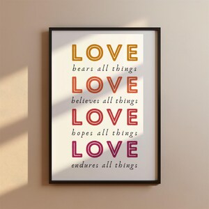 May include: A white framed print with the word "LOVE" repeated four times in different colors, each with a phrase below it. The colors are orange, red-orange, red, and pink. The phrases are "bears all things", "believes all things", "hopes all things", and "endures all things".