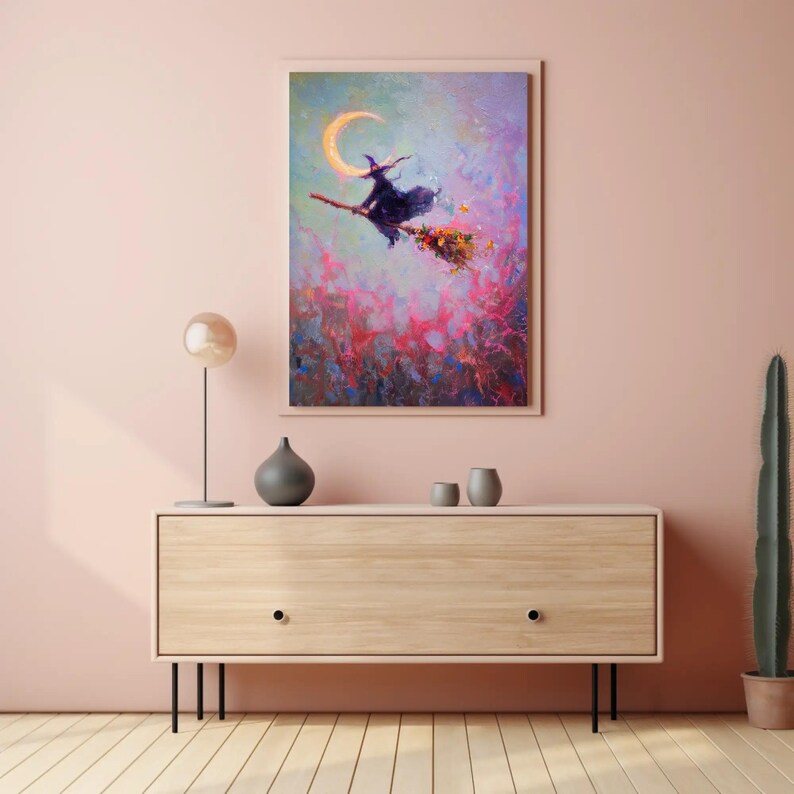 May include: A framed painting depicts a witch on a broomstick flying past a crescent moon. The witch is wearing a black hat and cloak. The background features a colorful, abstract design with pink, blue, and purple hues. The artwork is displayed above a wooden cabinet.