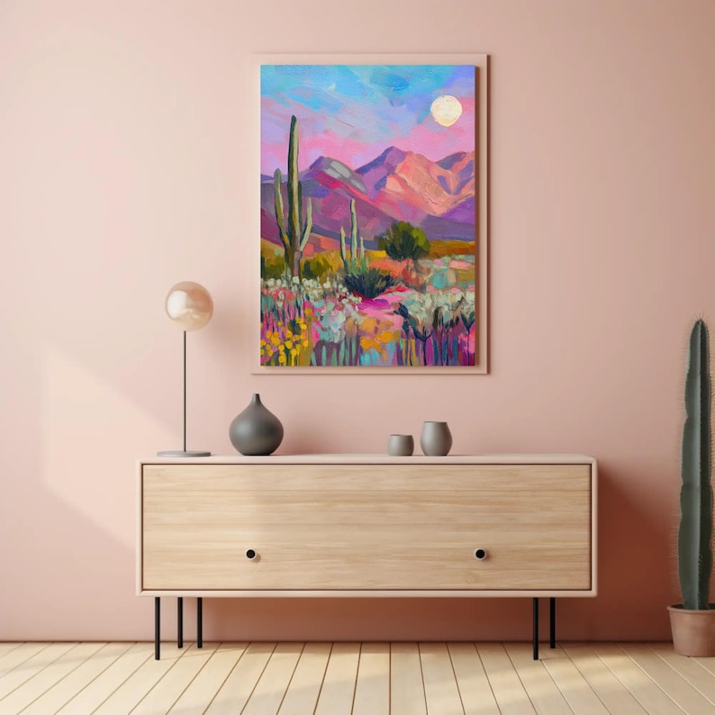 May include: An abstract landscape painting featuring a desert scene with cacti, mountains, and a full moon. The artwork uses vibrant colors like pink, purple, and blue. The painting is framed and displayed on a light wood cabinet.