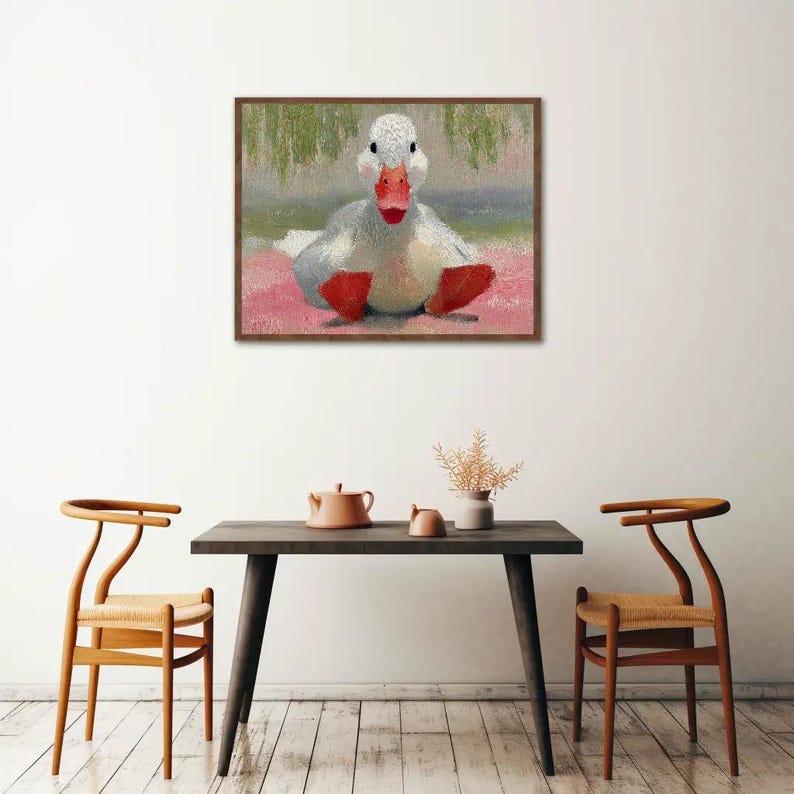 Puede incluir: Una obra de arte enmarcada representa un pato blanco con patas y pico rojos, sentado sobre una superficie rosa. El fondo presenta pinceladas verdes y rosas. La obra est&aacute; sobre una mesa de madera oscura con dos sillas.