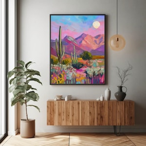 May include: A framed desert landscape painting with a vibrant color palette. The artwork depicts a desert scene with cacti, mountains, and a full moon. The colors include pinks, purples, blues, and yellows, creating a vivid and eye-catching piece.