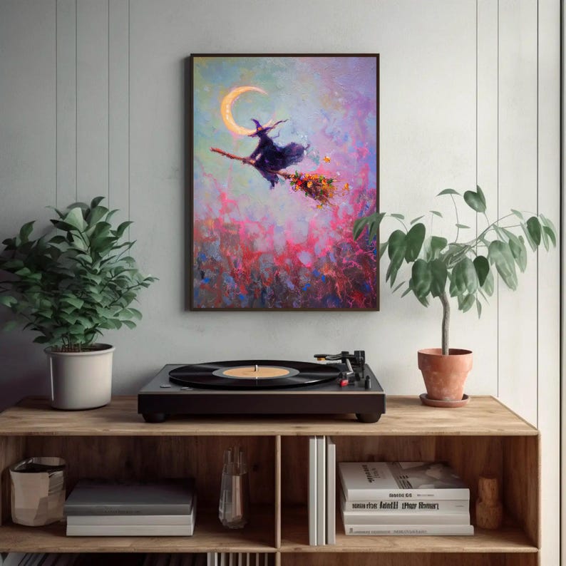 May include: A framed painting depicts a witch on a broomstick flying past a crescent moon. The artwork features a vibrant color palette of pinks, purples, and blues. The painting is displayed above a record player on a wooden shelf, with potted plants on either side.