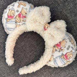 May include: White fuzzy headband with Winnie the Pooh ears. The ears are made of white fabric with a Winnie the Pooh print featuring Winnie the Pooh and Piglet.