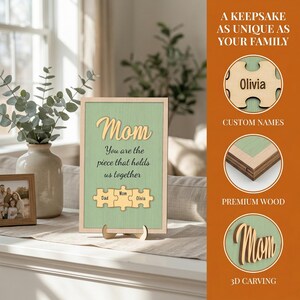 K&ouml;nnte beinhalten: Holzschild mit dem Wort "Mom" in Schreibschrift, vor hellgr&uuml;nem Hintergrund. Darunter steht der Text "You are the piece that holds us together". Enth&auml;lt Puzzleteile mit Namen. Das Schild wird auf einem Holzst&auml;nder pr&auml;sentiert.