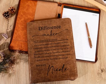 May include: A brown leather portfolio with the words "Difference Maker" and the name "Nicole" engraved on the front. The portfolio is open, revealing a notepad and pen. The open portfolio is on a wooden surface.