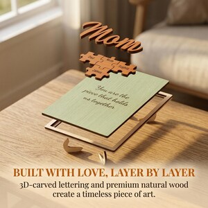 K&ouml;nnte beinhalten: Ein Holzschild mit der Aufschrift &bdquo;Mom&ldquo; und Puzzleteile mit Namen schweben &uuml;ber einem Holzrahmen mit einem gr&uuml;nen Einsatz. Der Einsatz lautet: &bdquo;Du bist das Teil, das uns zusammenh&auml;lt.&ldquo; Der Satz &bdquo;BUILT WITH LOVE, LAYER BY LAYER&ldquo; steht unten.