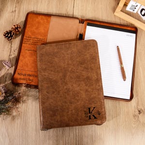 Op de afbeelding: Een bruine leren portfolio met ritssluiting, open om een notitieblok en pen te laten zien. De omslag is in reliëf voorzien van de letter "K" en een bloemenontwerp. De binnenkant heeft de tekst "NOAH SMITH MARKETING".