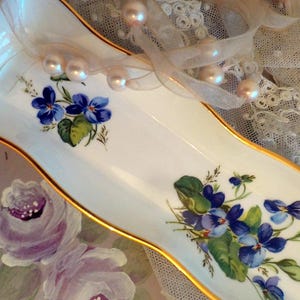 May include: An elongated white porcelain tray with a gold-trimmed rim, featuring two distinct hand-painted clusters of blue violet flowers with green leaves. The tray has a curved, wave-like shape and a smooth, glossy finish on its surface.