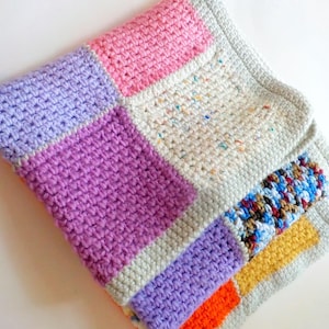 May include: A handmade crochet blanket with a patchwork design. The blanket features squares of pink, purple, and orange, with a speckled white square. The border is a light grey colour, and the blanket is folded.
