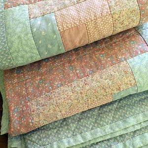 May include: A folded patchwork quilt featuring a variety of floral and solid fabric squares in shades of sage green, dusty rose, and cream. The textile displays intricate diamond-patterned machine quilting throughout the surface of the fabric.
