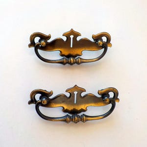May include: Two matching brass-toned metal drawer pulls featuring a decorative backplate with a central keyhole cutout and a curved, swinging bail handle. The hardware has a dark, aged finish with smooth, symmetrical scrollwork details.