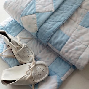 May include: A folded quilt featuring a chequered pattern of light blue and white squares with subtle polka dots, paired with a pair of white leather moccasin-style shoes that have lace-up closures and decorative perforated detailing on the toe.