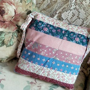 Peut inclure: Un coussin carré matelassé avec un motif patchwork. Il présente des rayures horizontales de tissus floraux roses, bleus et blancs. Le coussin a une bordure bordeaux et une sangle en tissu assortie. Mesure environ 30 cm x 30 cm.