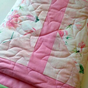 May include: A folded quilt featuring a patchwork design of solid pink fabric and a white fabric printed with pink roses and green leaves. The textile is finished with decorative curved stitching and a solid light green border along the bottom edge.