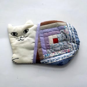 Peut inclure: Une pochette faite à la main en forme de chat. Le visage du chat est blanc avec des détails noirs brodés. La section de la pochette présente un motif patchwork avec divers tissus dans les tons de bleu, violet, marron et blanc. La pochette est probablement utilisée pour de petits objets.
