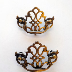 May include: Two antique brass drawer pulls with an ornate, openwork design. Each pull features a decorative centre and curved handles with small ball accents. The pulls are set against a white background.
