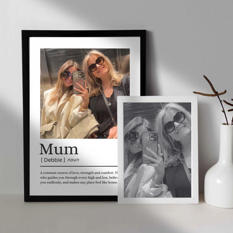 May include: Two framed black and white photos of a mother and daughter taking a selfie. The larger frame has the word "Mum" and a definition below it. A white vase with a plant is to the right.