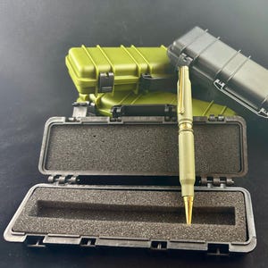 May include: A green pen in a black and green hard case. The pen is made from a bullet casing and has a gold tip.