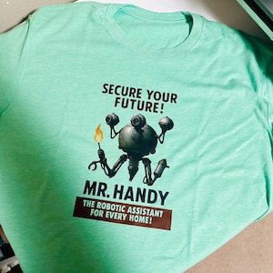May include: A light green t-shirt featuring a graphic of a robot with the text "SECURE YOUR FUTURE!" above and "MR. HANDY THE ROBOTIC ASSISTANT FOR EVERY HOME!" below. The robot has multiple arms and cameras.