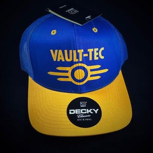 May include: A blue and yellow baseball cap with a black logo that says "Vault-Tec" and a yellow Vault-Tec logo. The cap is made by "Decky Classic Original".