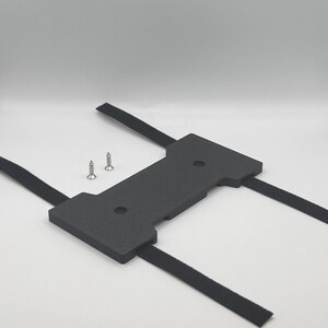 May include: A black, rectangular component with two screws and black straps. The component has a central section with two holes and two protruding sections. The black straps are attached to the component.