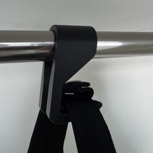 Może przedstawiać: Czarny, kątowy wspornik jest przymocowany do srebrnego metalowego pręta. Czarny pasek jest połączony ze wspornikiem. Wspornik wydaje się być wykonany z tworzywa sztucznego. Tło jest białe.