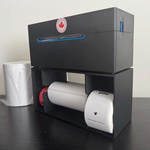 May include: Black label printer with a blue trim and a Canadian flag sticker. Rolls of white labels are visible, including one with a heart and the text "Thank you. I hope you have a good time."