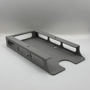 Pode incluir: Um suporte de produto retangular cinza com interior rebaixado e bordas arredondadas. O suporte possui vários recortes retangulares nas laterais e um recorte curvo em uma extremidade. O produto é feito de um material sólido.