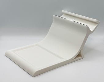 Puede incluir: Un soporte curvo blanco de cerámica para teléfono, con una base rectangular y una sección superior elevada. El soporte tiene un diseño moderno y minimalista, adecuado para sostener un teléfono o tableta. El soporte es de material liso.