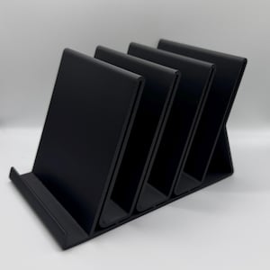 Puede incluir: Un organizador de escritorio negro con cuatro compartimentos angulados. El organizador está hecho de un material sólido y tiene un diseño simple y moderno. Está diseñado para guardar papeles, archivos u otros suministros de oficina. El organizador se muestra sobre un fondo blanco.