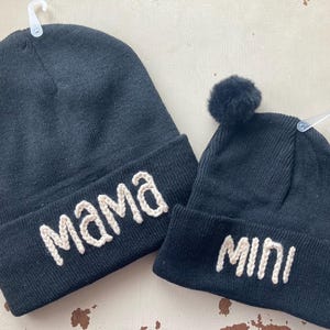 May include: Two black knit beanies with white embroidered text. The larger beanie says "Mama" and the smaller beanie says "Mini".