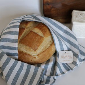 May include: A round loaf of bread wrapped in a blue and white striped cloth. The cloth has a tag that reads "HEARTH AND HEM". The bread is golden brown with a scored top. A wooden cutting board and a small container are in the background.
