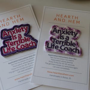 May include: Two iron-on patches with the text "Anxiety is a Terrible Life Coach" in purple and pink embroidery. The patches are on white cards with the brand name "HEARTH AND HEM".