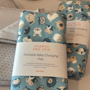 May include: A teal portable baby changing pad with a pattern of cartoon animals, including owls, cows, and bunnies. The pad is labeled "HEARTH AND HEM" and "Portable Baby Changing Pad". The text also mentions a waterproof PUL lining.
