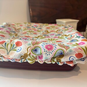 May include: A burgundy rectangular baking dish covered with a floral patterned fabric lid. The lid features a white background with pink, orange, blue, and green floral and leopard print designs. The dish is on a white surface.