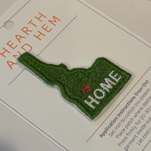 May include: Green embroidered patch in the shape of the state of Idaho, with the word "HOME" in white. A small red heart is stitched near the center. The patch is on a white card with the words "HEARTH AND HEM".