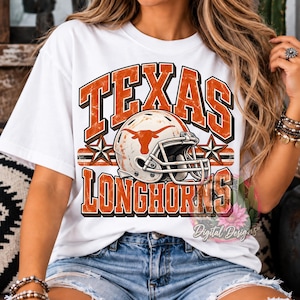 Può includere: Una t-shirt bianca con una stampa grafica che riporta il testo TEXAS LONGHORNS in lettere arancioni dall'effetto usurato. Il design include un casco da football americano bianco con un logo longhorn arancione, affiancato da due stelle su una riga.