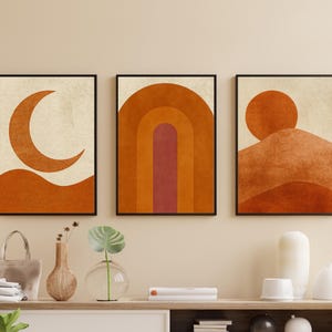 May include: Set of three framed abstract art prints. Each print features a minimalist design in shades of orange and beige. One depicts a crescent moon, another a layered arch, and the third a sun over a mountain.