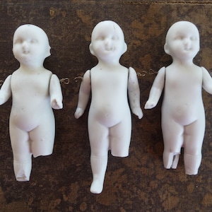 May include: Three antique bisque porcelain doll parts. Each doll has a smiling face, arms outstretched, and legs. The dolls are white and have some damage. The dolls are connected by a thin wire.
