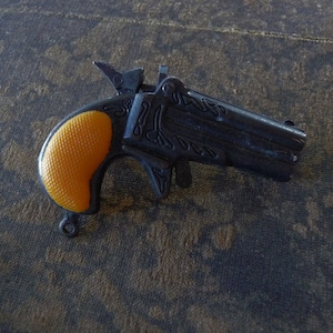 May include: A small, black toy gun with an orange plastic handle. The gun has intricate details and a decorative design.